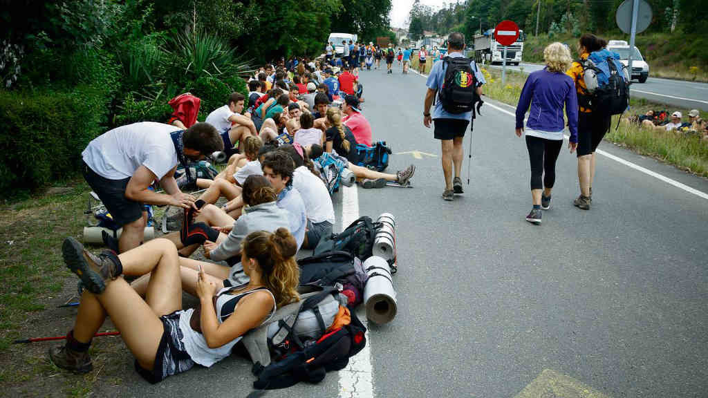 Grupo numeroso de peregrinas e peregrinos do Camiño de Santiago descansando ao carón dunha estrada (Foto: @hosvidalet).