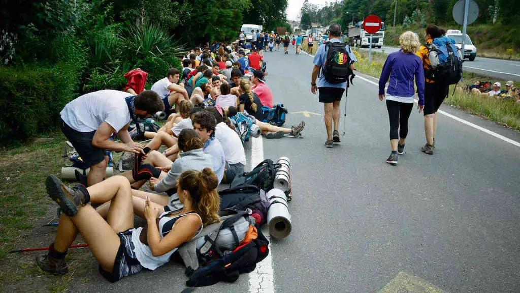 Grupo numeroso de peregrinas e peregrinos do Cami&ntilde;o de Santiago descansando ao car&oacute;n dunha estrada (Foto: @hosvidalet).